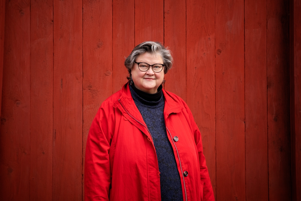 Marikki Hakola seisoo, hymyilee ja katsoo suoraan kameraan.. Taustalla punainen seinä.
